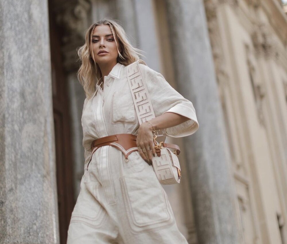 Woman wearing a cream-colored outfit with a belt and crossbody bag, posing between classic architectural columns.
